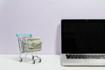 macbook pro on white table beside a miniature shopping cart with money