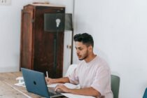 Young ethnic man working on laptop and writing in copybook