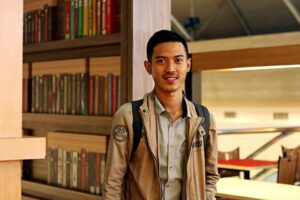 Man in Brown Jacket Standing Near Wooden Book Shelves