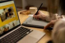 A young woman is sitting at a desk in her home, working on a laptop and using a tablet.