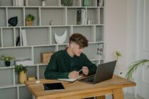 A Male Student in Green Long Sleeves Using a Laptop on a Wooden Desk