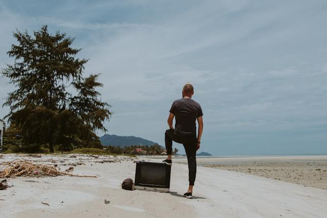 back view of a man standing on the beach and resting his foot on an old tv