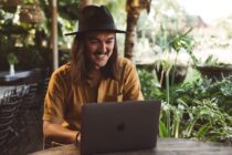 portrait of a smiling man with long hair and hat using computer in a garden