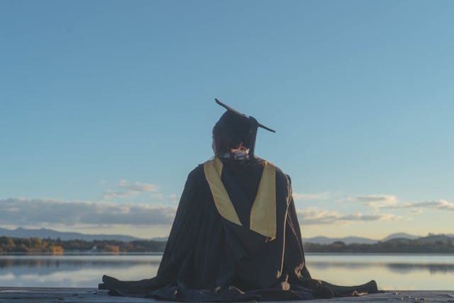 back view of a student wearing a graduation gown and a mortarboard sitting by a lake
