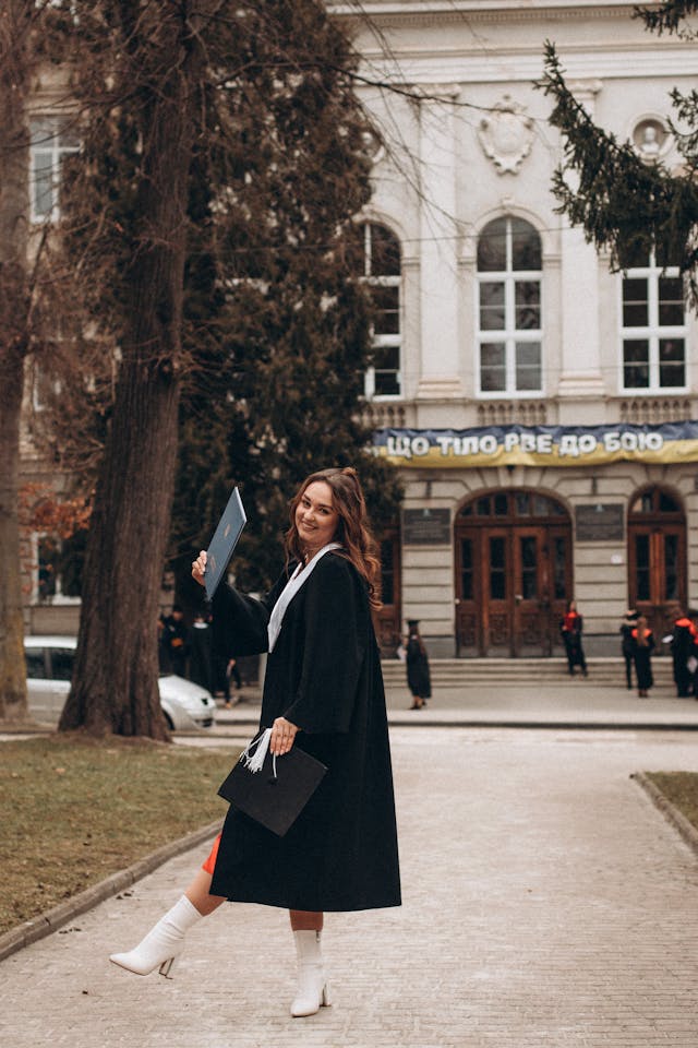 brunette woman in graduation gown holding cap and diploma