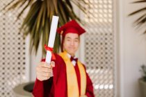 a man wearing red toga and graduation cap holding a rolled certificate while smiling at the camera